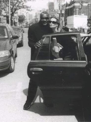 Two stylish individuals pose by a car on an urban street, captured in black and white.