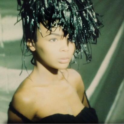Model with large teased black hair, wearing a strapless black top, photographed with dramatic lighting.