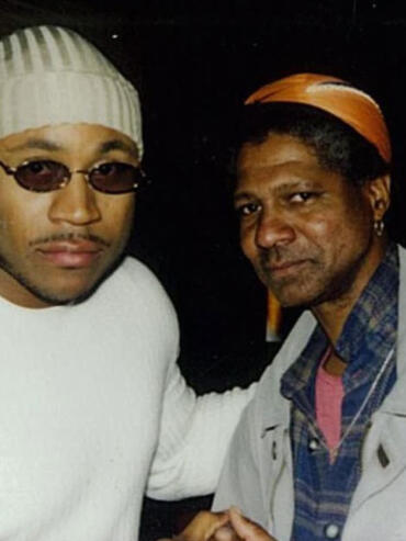 Two men pose closely, one wearing sunglasses and a striped beanie, the other with an orange headscarf and checked shirt.