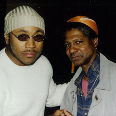 Two men pose closely, one wearing sunglasses and a striped beanie, the other with an orange headscarf and checked shirt.