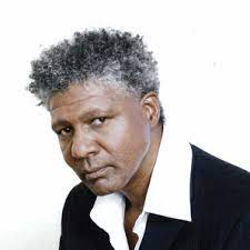 Close-up of a man with short, curly gray hair wearing a black blazer over a white shirt.