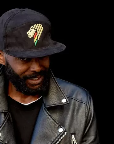 Man wearing a black cap with colorful emblem and a black leather jacket.