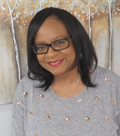Woman with shoulder-length black hair, glasses, red lipstick, wearing a gray sweater with gold embellishments.