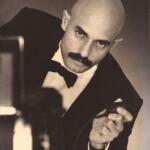Black-and-white image of a man with a bald head and mustache, dressed in formal attire with a bow tie, holding a vintage microphone and looking directly at the camera.
