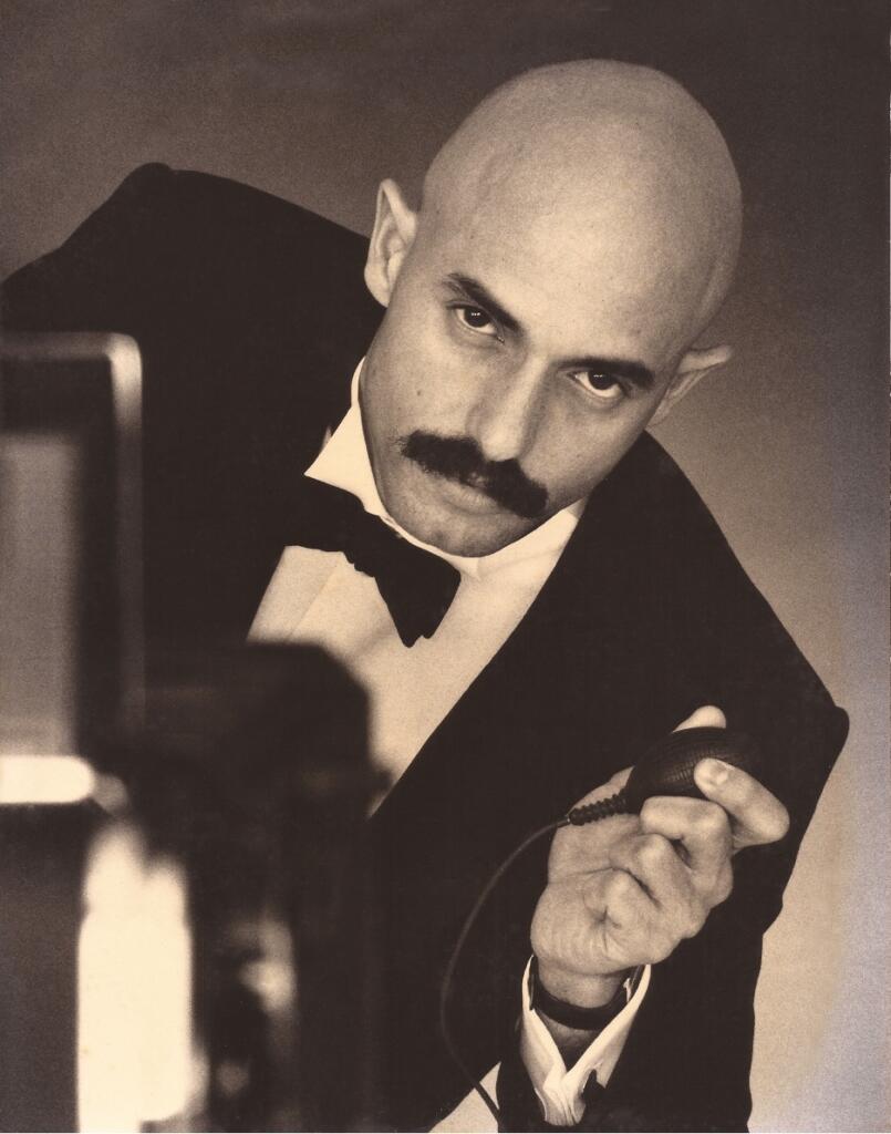 Black-and-white image of a man with a bald head and mustache, dressed in formal attire with a bow tie, holding a vintage microphone and looking directly at the camera.