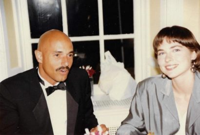 A man in a tuxedo and a woman with short hair in a gray blazer smiling indoors.