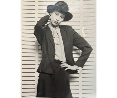 Black-and-white image of a woman in vintage fashion, wearing a tailored blazer, blouse, and a stylish hat.