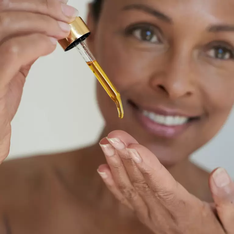 A woman applying skincare serum from a dropper with a bright smile.