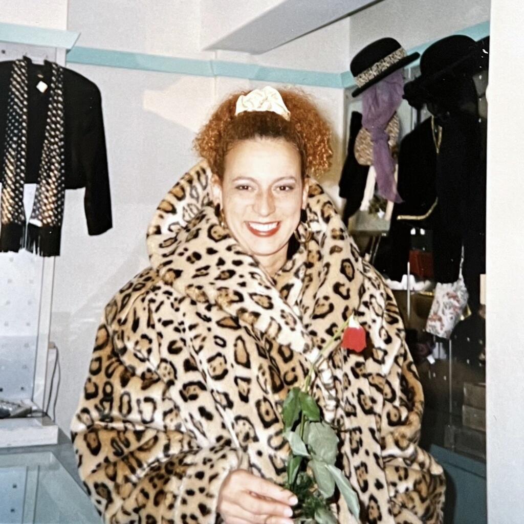 Woman in leopard-patterned coat holding a red rose, smiling, with a vintage clothing display in the background.