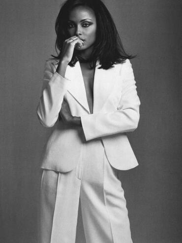 Black and white portrait of a woman in a tailored suit with one hand near her face, exuding confidence.