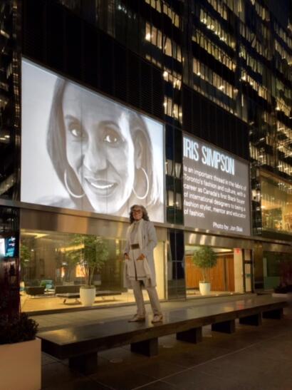 A woman in stylish beige ensemble stands outside a modern city building at night, illuminated by large digital displays.