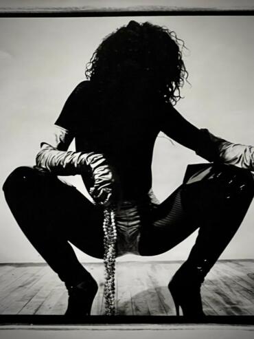 A silhouette of a woman with curly hair, wearing a fitted jacket, thigh-high boots, and holding a beaded accessory, pose in front of a minimal background.