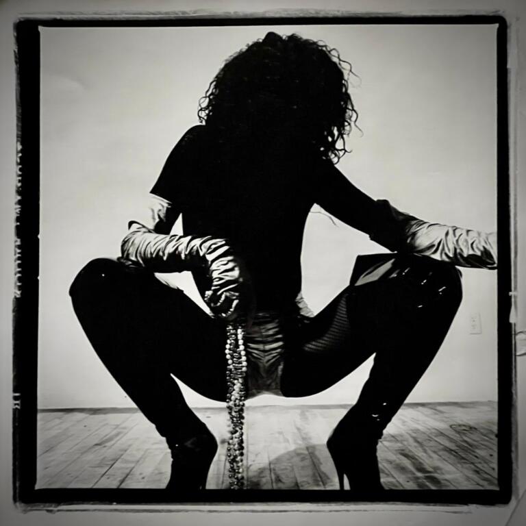 A silhouette of a woman with curly hair, wearing a fitted jacket, thigh-high boots, and holding a beaded accessory, pose in front of a minimal background.
