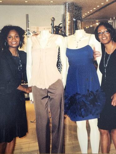 Two women stand beside mannequins dressed in elegant garments at an indoor fashion event.