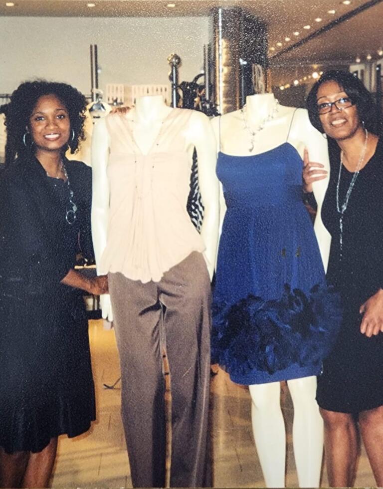 Two women stand beside mannequins dressed in elegant garments at an indoor fashion event.