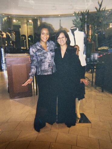 Two women posing together in a stylish indoor setting, dressed in elegant black and patterned outfits.