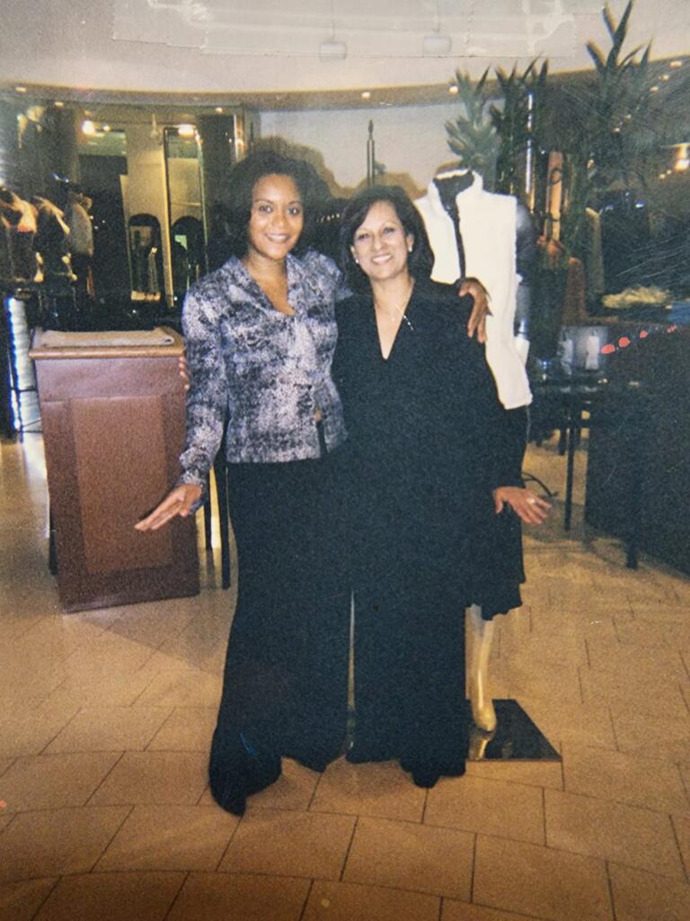Two women posing together in a stylish indoor setting, dressed in elegant black and patterned outfits.