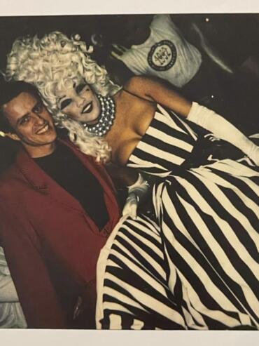 A person dressed in a striped black-and-white gown with platinum blonde curls poses with a man in a red blazer.