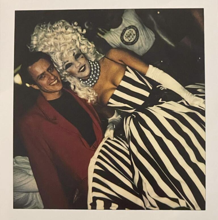 A person dressed in a striped black-and-white gown with platinum blonde curls poses with a man in a red blazer.