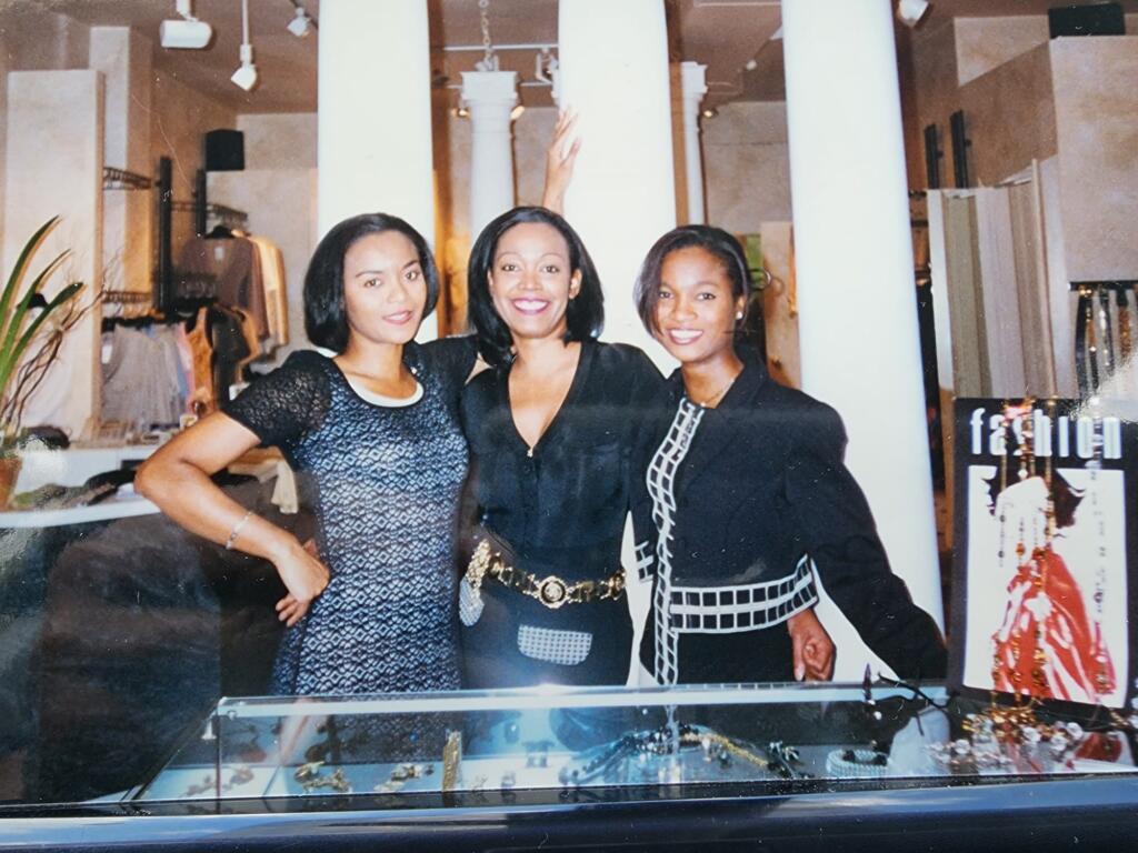 Three women standing together inside a boutique store, smiling and dressed in stylish black outfits.
