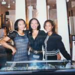 Three women standing together inside a boutique store, smiling and dressed in stylish black outfits.