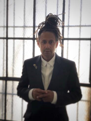Person with dreadlocks in a blazer and white shirt, standing against a textured glass window.
