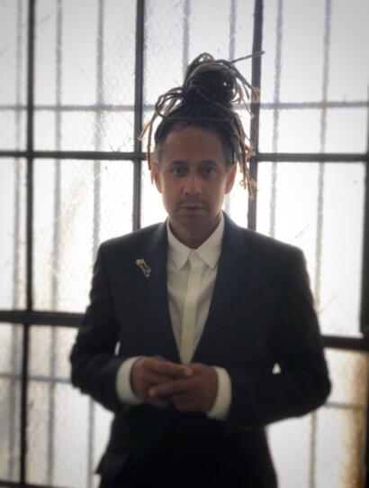 Person with dreadlocks in a blazer and white shirt, standing against a textured glass window.