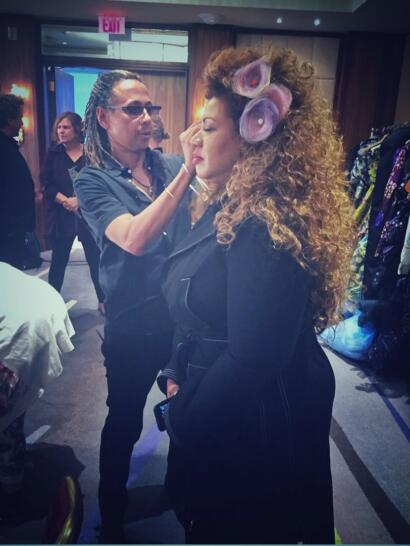 Makeup artist applying finishing touches to a woman with voluminous curly hair and floral hair accessories.