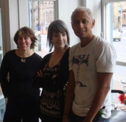 Group of three casually dressed people standing near a window in a brightly lit indoor space.