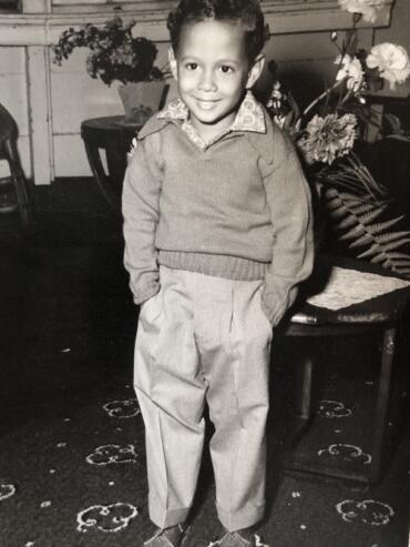 A young boy in a sweater and pleated trousers, smiling indoors with flowers in the background.