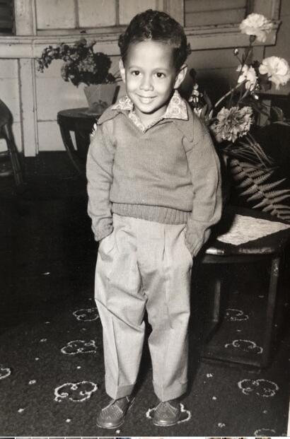 A young boy in a sweater and pleated trousers, smiling indoors with flowers in the background.
