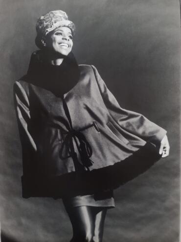 Black and white portrait of a woman smiling, wearing a hat, scarf, and stylish coat with puffed sleeves.