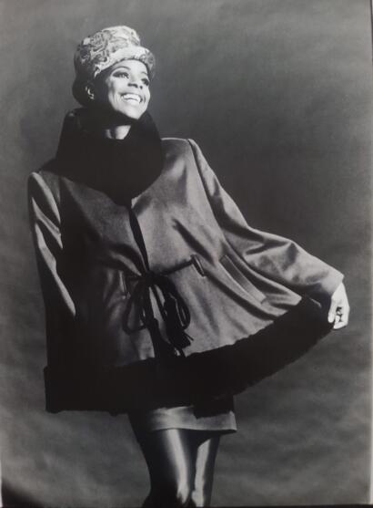 Black and white portrait of a woman smiling, wearing a hat, scarf, and stylish coat with puffed sleeves.