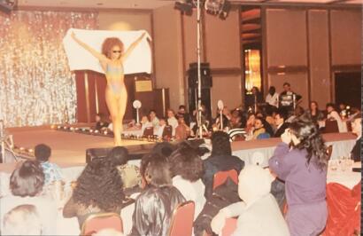A model in colorful swimwear strikes a pose on the runway during a fashion show, with an audience watching.
