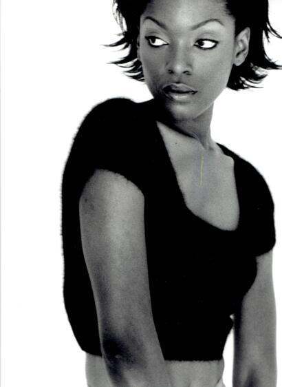 Black-and-white portrait of a woman with short, textured hair, wearing a black top and a delicate necklace, posed with a confident expression.