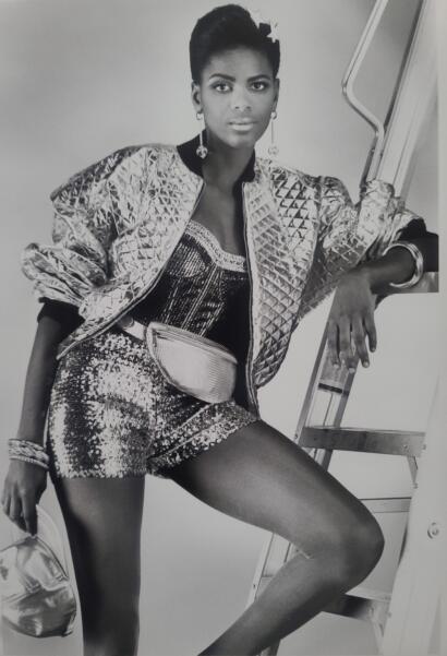 Black-and-white fashion photo of a woman in a sequin top, quilted jacket, and shorts, posed against a ladder.