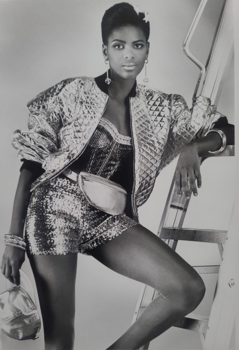 Black-and-white fashion photo of a woman in a sequin top, quilted jacket, and shorts, posed against a ladder.