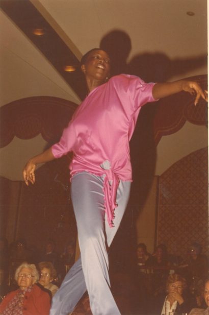 Woman in a pink satin blouse and light gray pants dancing in front of an audience.