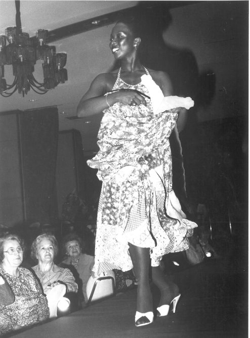 Black-and-white photo of a woman confidently walking on a runway in a floral, flowing dress and high heels, with an attentive audience watching.