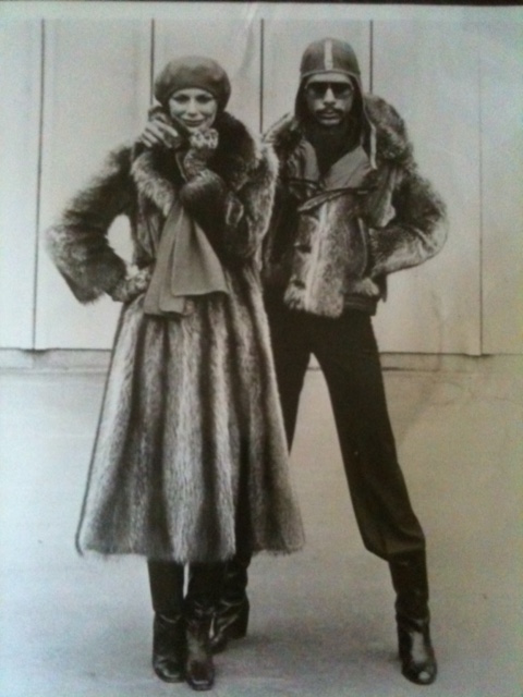 Black-and-white photo of a woman in a fur coat and hat, and a man in a leather jacket with fur accents, both dressed in vintage winter fashion.