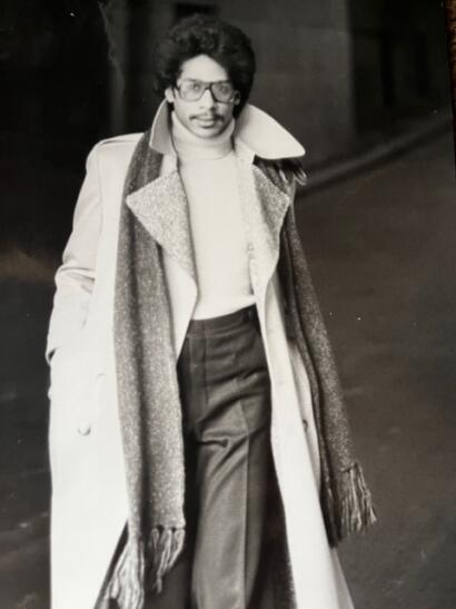 Black-and-white photo of a stylish woman wearing a long coat, high-waisted pants, turtleneck, glasses, and a scarf, captured outdoors.