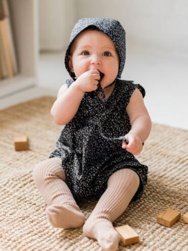 Adorable baby girl in a black floral hooded dress, sitting on a woven rug with wooden blocks.