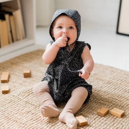 Adorable baby girl in a black floral hooded dress, sitting on a woven rug with wooden blocks.