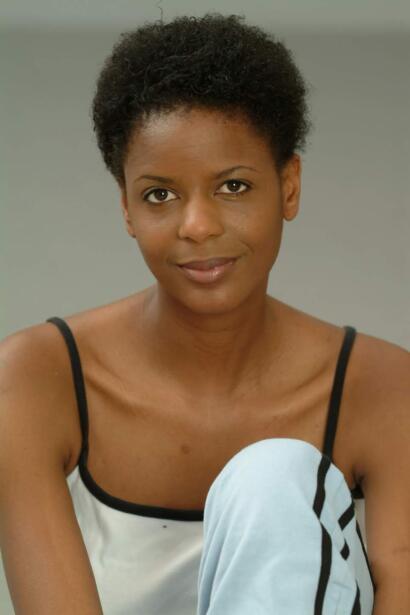 Portrait of a woman with short natural hair, wearing a white top with black straps, against a neutral background.