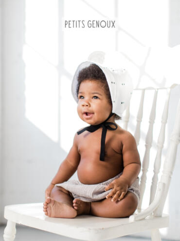 Adorable baby with a bonnet, sitting shirtless on a white wooden chair in a softly lit space.