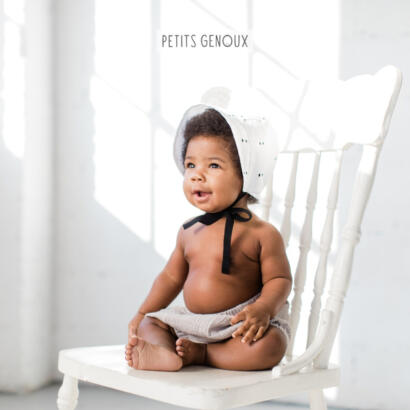 Adorable baby with a bonnet, sitting shirtless on a white wooden chair in a softly lit space.