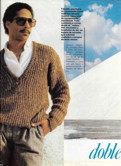 Man wearing a white shirt, brown sweater, grey pants, and sunglasses against a coastal background.