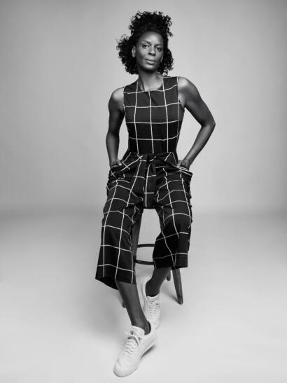 Black-and-white portrait of a woman in a plaid jumpsuit with sneakers, sitting on a stool.