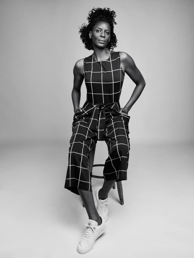 Black-and-white portrait of a woman in a plaid jumpsuit with sneakers, sitting on a stool.