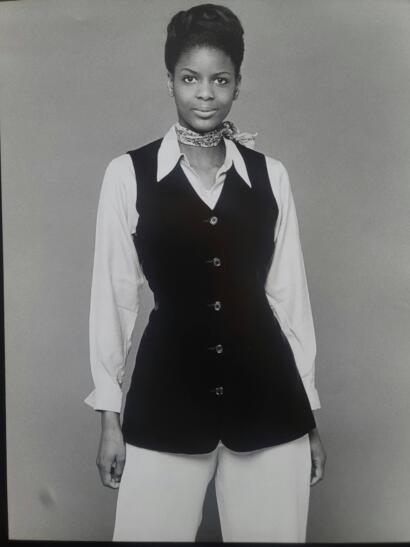 Black-and-white portrait of a woman wearing a collared shirt, a fitted vest, and a scarf around her neck, captured in a fashion magazine style.
